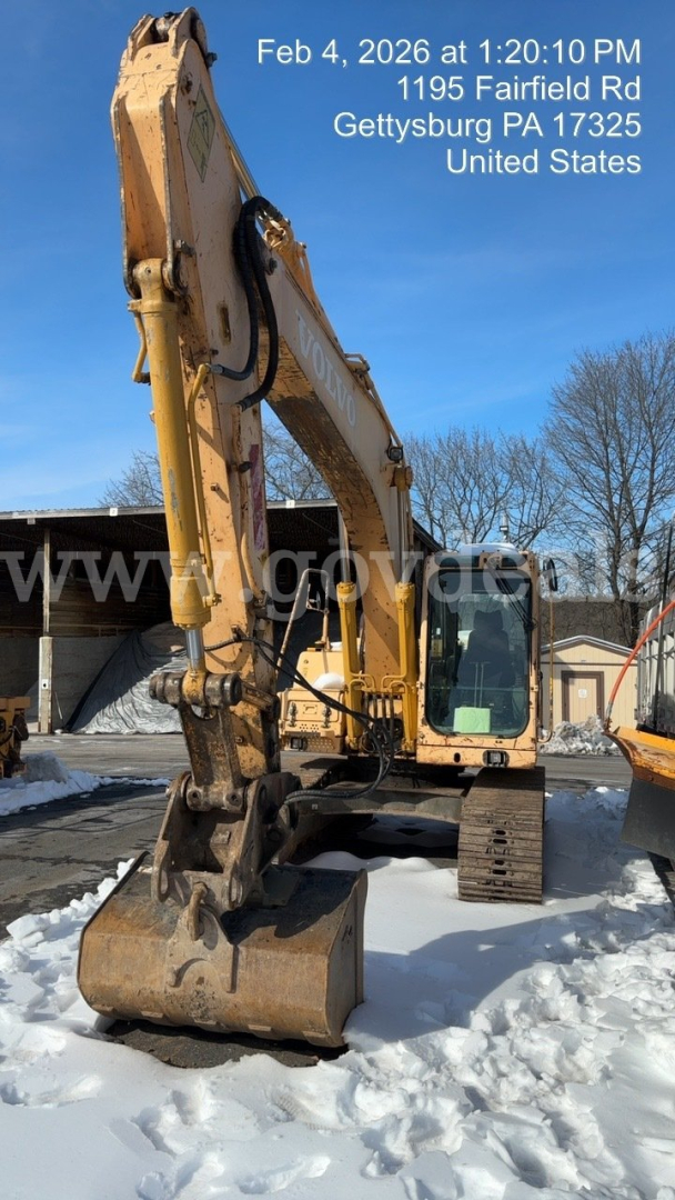 Volvo EC160B excavator in side view with bucket on ground, snow on the ground, blue sky, watermarked text with date and address.