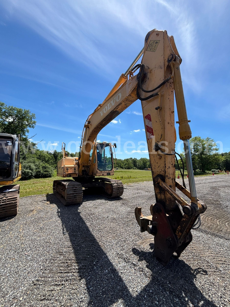 Volvo EC160B excavator in service, orange hydraulic arm with bucket attachment, long reach boom, tracks on gravel surface, clear blue sky in background.