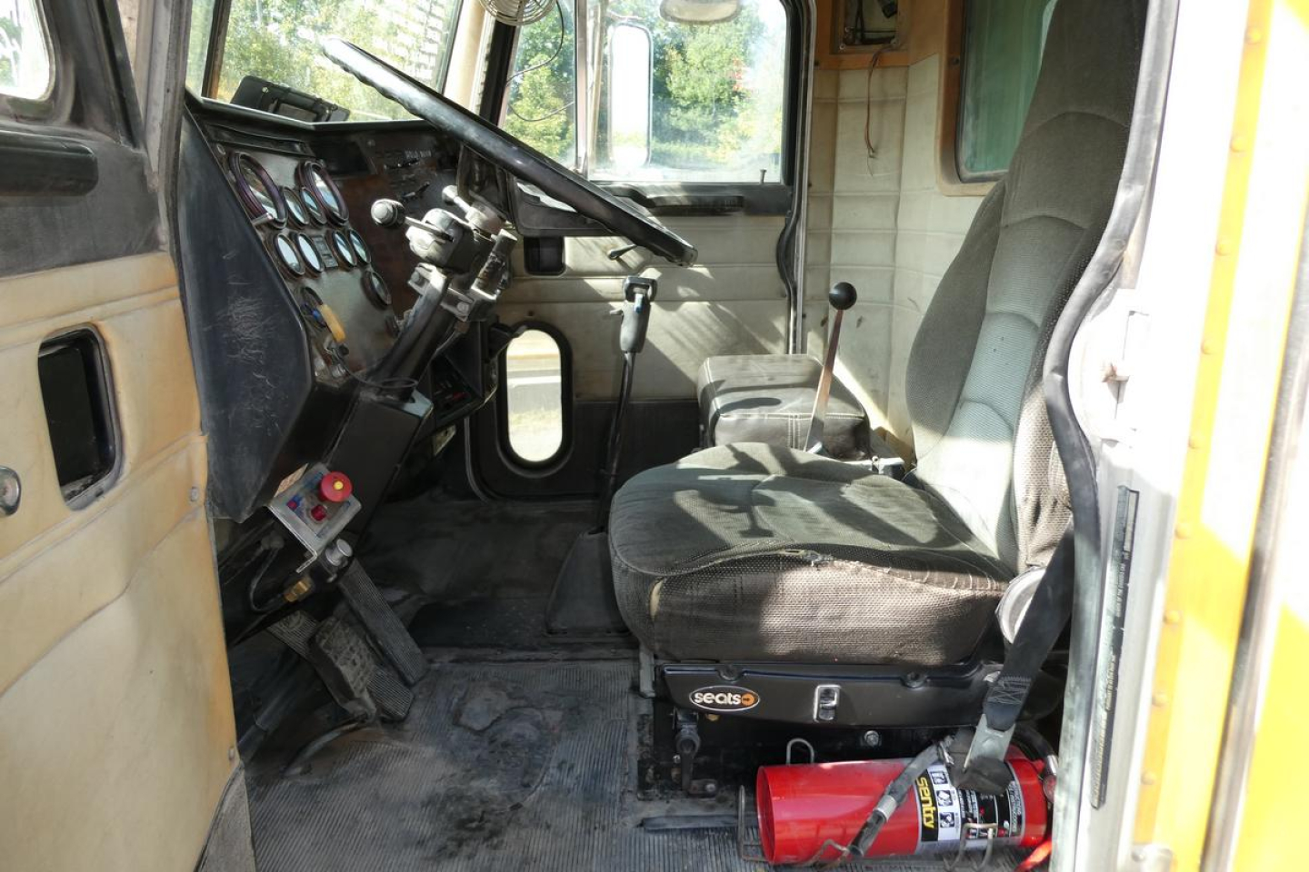 Truck cab interior showing driver seat and steering wheel with dashboard gauges; visible labels include a red fire extinguisher and a control panel.