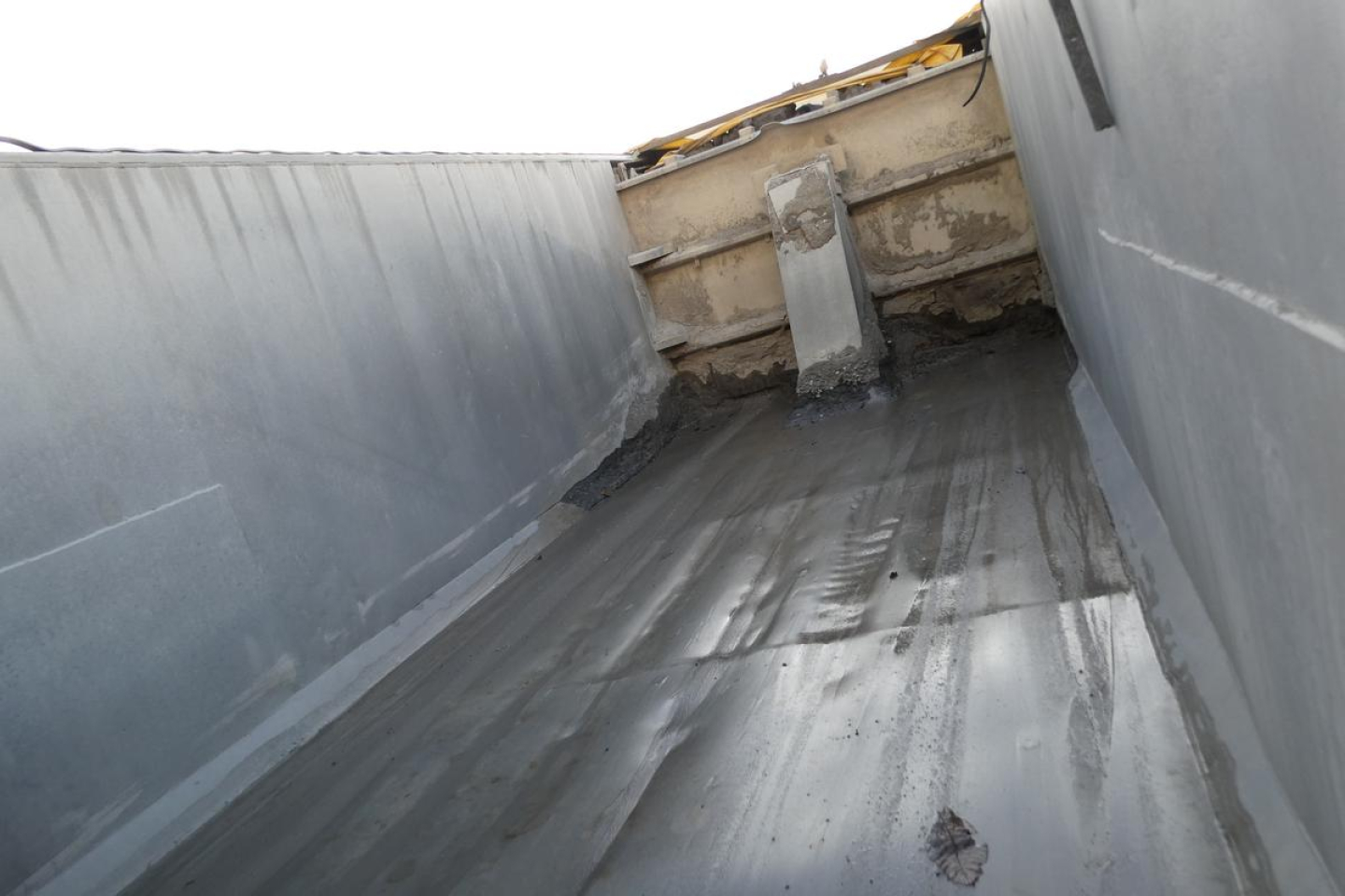 Dump truck bed interior with metal walls and a central chute, showing a weathered surface and drainage channel.