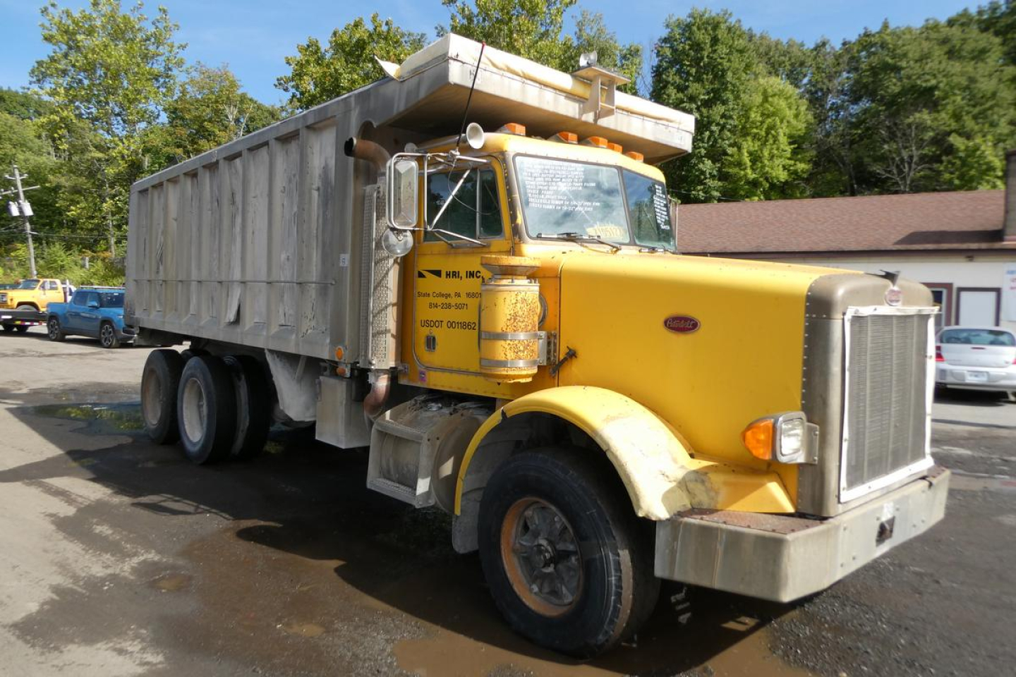 Peterbilt dump truck from a side angle showing yellow cab and gray dump body with chrome grille and bumper; visible text on the door and surroundings including company name and contact details.
