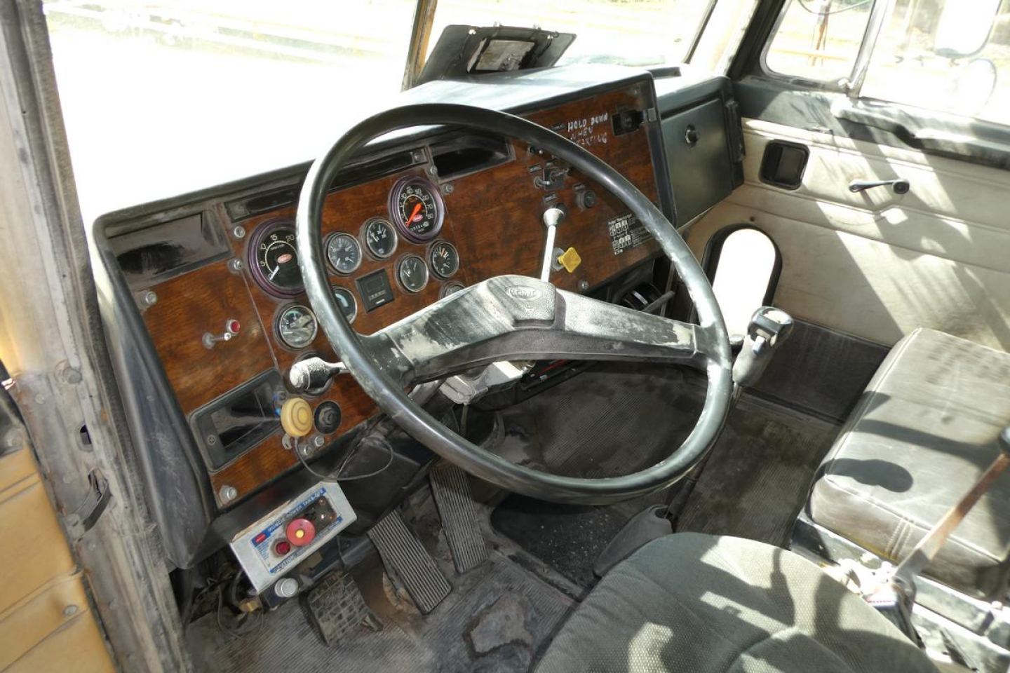 Peterbilt truck cab interior showing steering wheel, dashboard gauges, and wood-grain panel with switches and pedals.