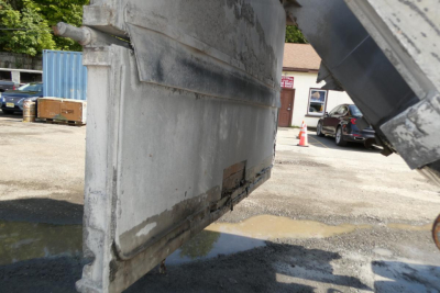 Dump truck bed side with metal hoist and chipped lower panel; wet ground and concrete barrier in background.