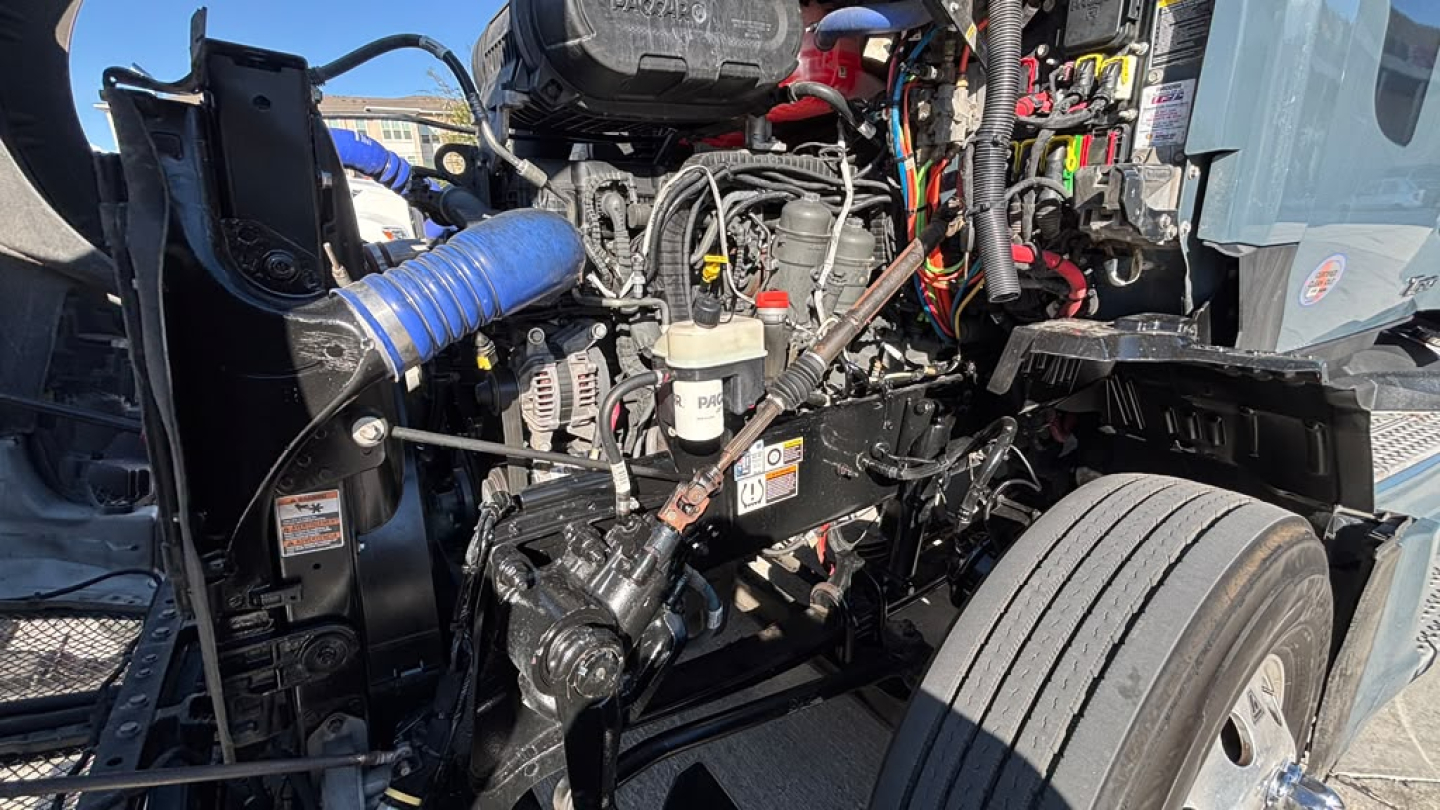 Kenworth T680 engine bay showing hydraulic lines, wiring loom, and dashboard components; visible components include hoses, alternator, belts, and steering linkage.