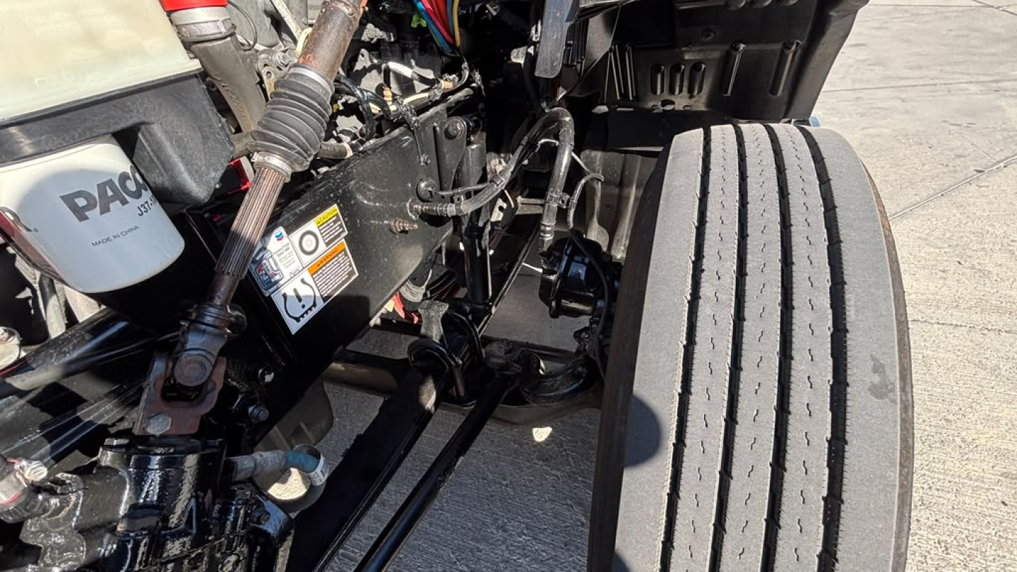 Kenworth T680 engine bay with front wheel and suspension components visible; various hoses and wires present.