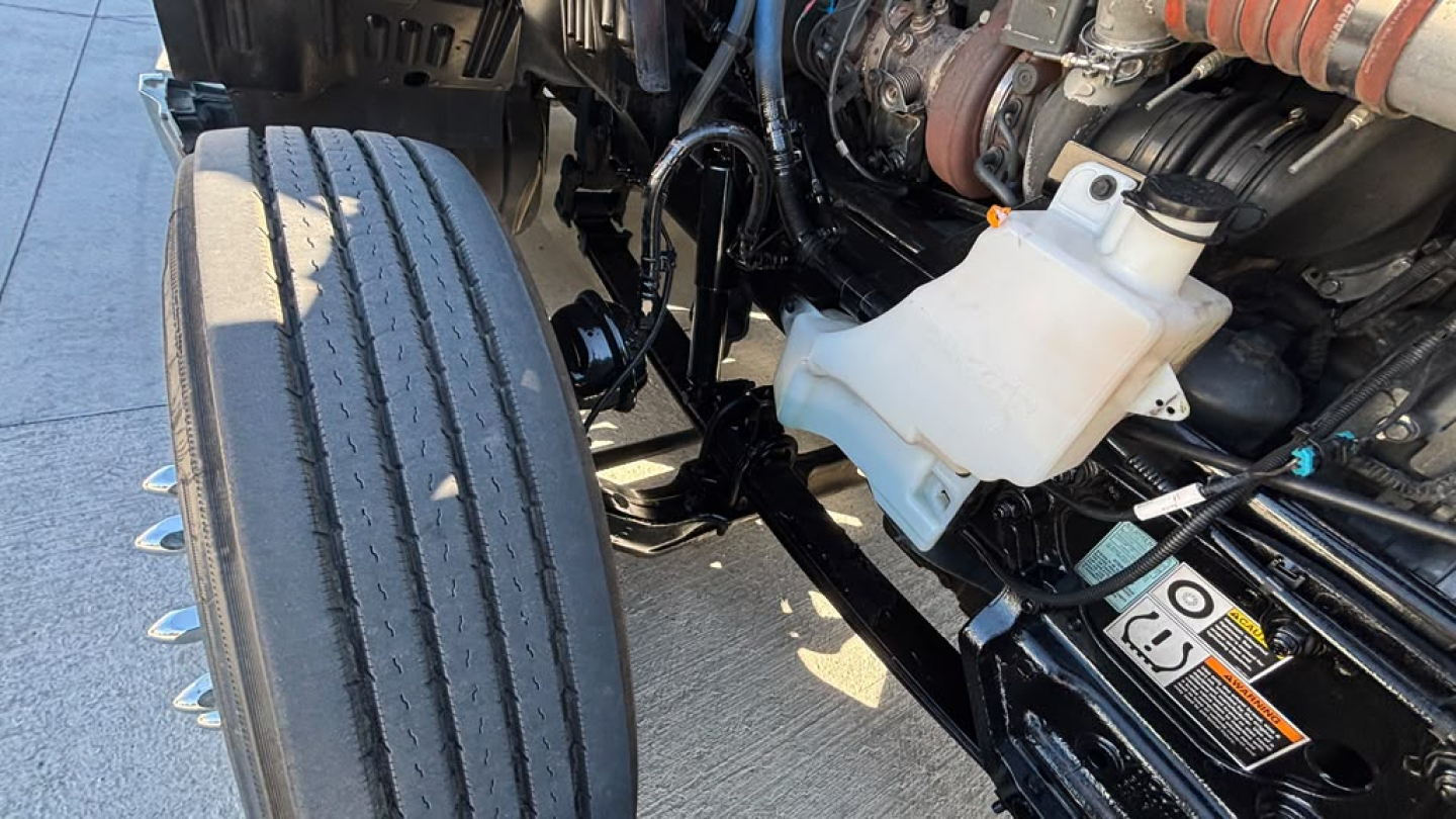 Kenworth T680 engine bay with front left dual tires visible, white coolant reservoir, hoses, and air intake; engine components and frame in view.