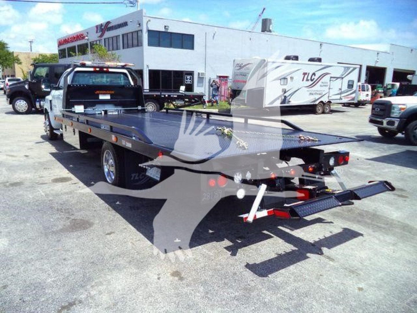 Flatbed tow truck on a dealership lot with a white building and other vehicles in the background, showing a 2023 Chevrolet Silverado 6500HD rollback truck.