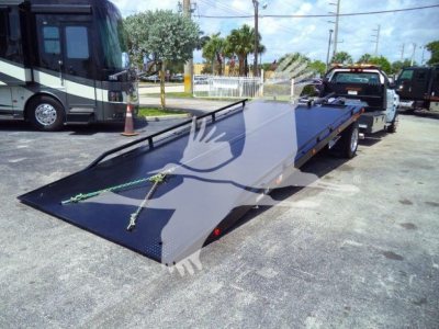 Flatbed tow truck in a dealership lot with another bus in background; contains a long black steel ramp with toe hooks and a green strap lying on it.