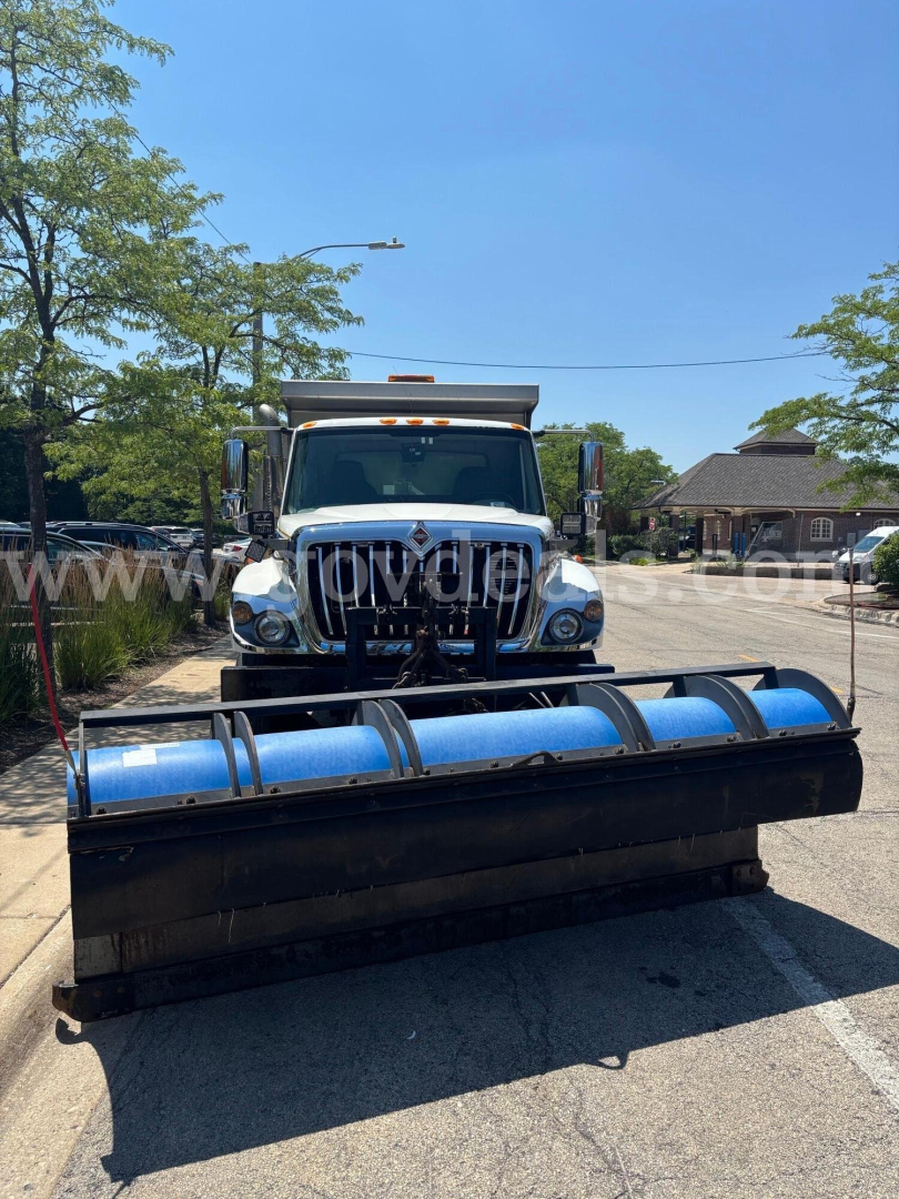 Front view of a 2009 International 7400 with a large blue snow plow blade attached. Vehicle has chrome grille, dual mirrors, and warning light bar on cab; no visible branding text legible besides truck features.
