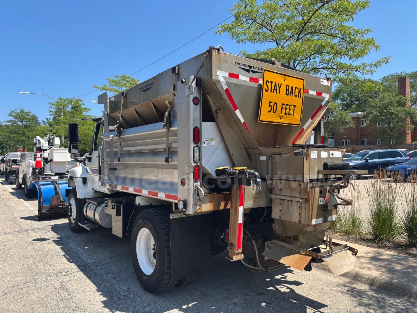 Dump truck shown from rear three-quarter view with amber warning sign Stay Back 50 Feet on tailgate and utility bed with hoses and rear hitch.
