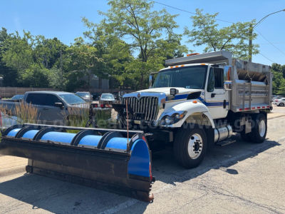 Heavy-duty International 7400 dump truck with a blue snow plow attachment in front, cab with white and blue livery, bed raised, side mirrors, and a boom on top.