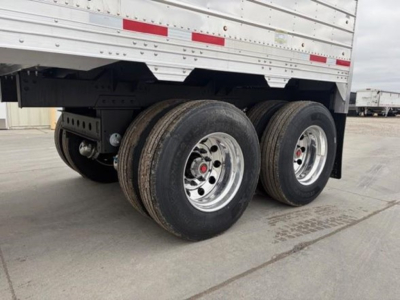 Trailer axle view showing dual rear axles and tires beneath a silver Timpte hopper trailer, with chrome rims and red hub caps.
