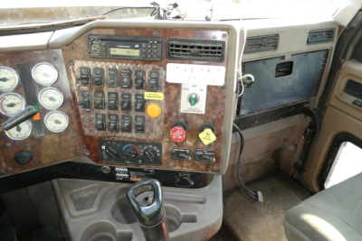 Truck cab interior panel with multiple gauges, switches, and a wood-grain dashboard; gear shifter visible in foreground.