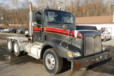International 9400 conventional daycab truck showing chrome exhaust stack, black cab with red and gold stripe decals, front grille, dual rear axles, and a rusted front bumper.