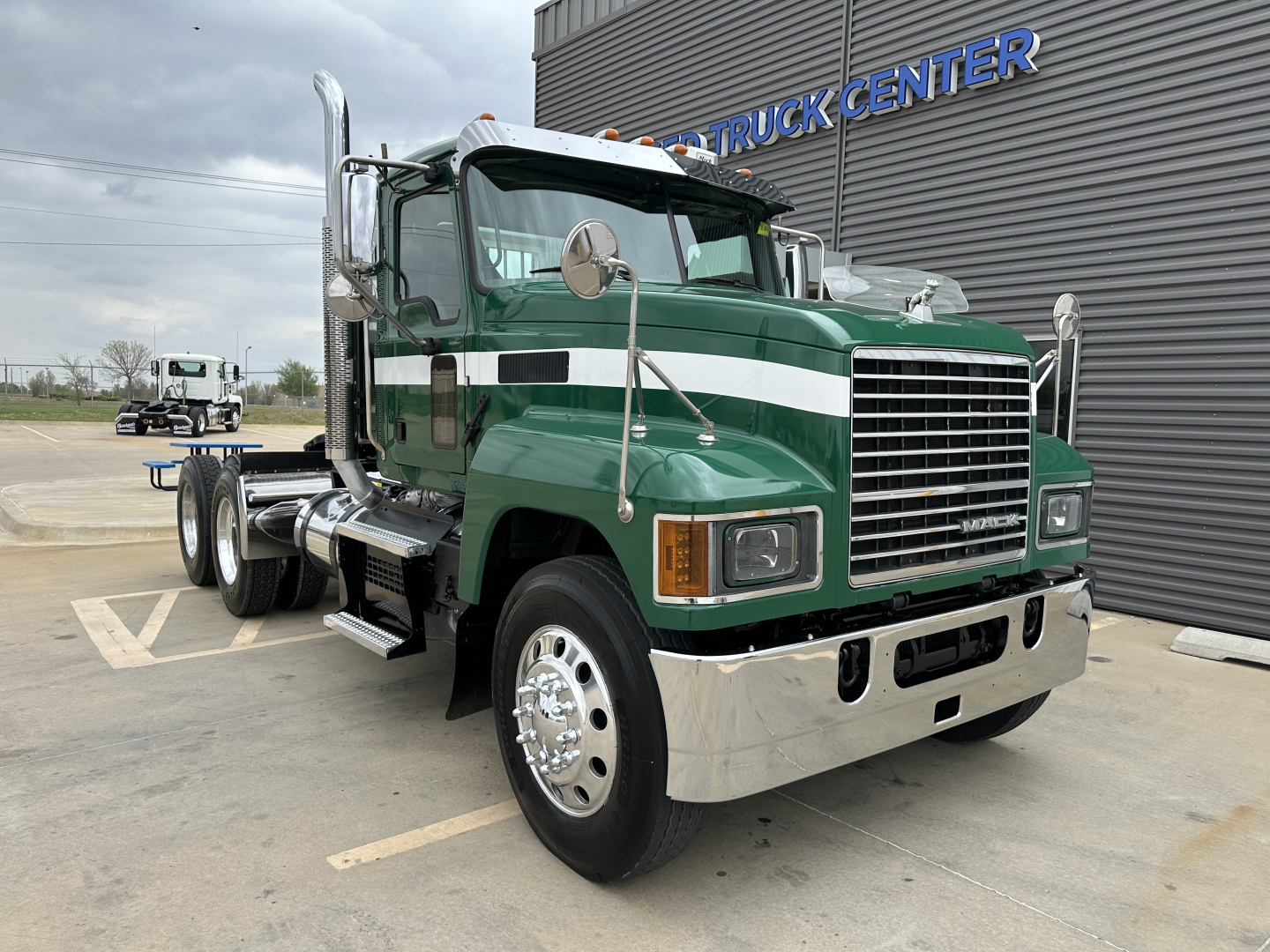 Mack PI64T conventional daycab truck in green with white stripe, front grille, large chrome bumper, dual exhaust stacks, parked outside dealership.