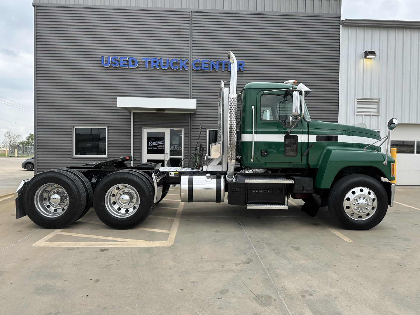 Mack PI64T conventional daycab truck side view showing green and white cab and dual rear axles in front of Used Truck Center building.