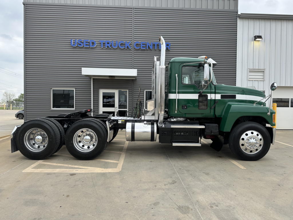 Mack PI64T conventional daycab truck side view showing green and white cab and dual rear axles in front of Used Truck Center building.