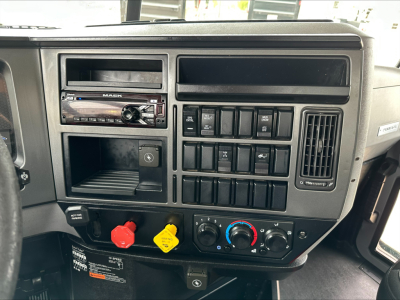 Cabin dashboard of a 2020 Mack PI64T showing radio, assorted switches, climate controls, red emergency brake knob, and yellow cap.