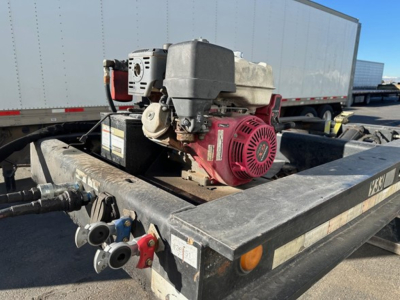 Lowboy trailer showing a small gasoline-powered engine/generator mounted on the frame with hoses and control valves visible.
