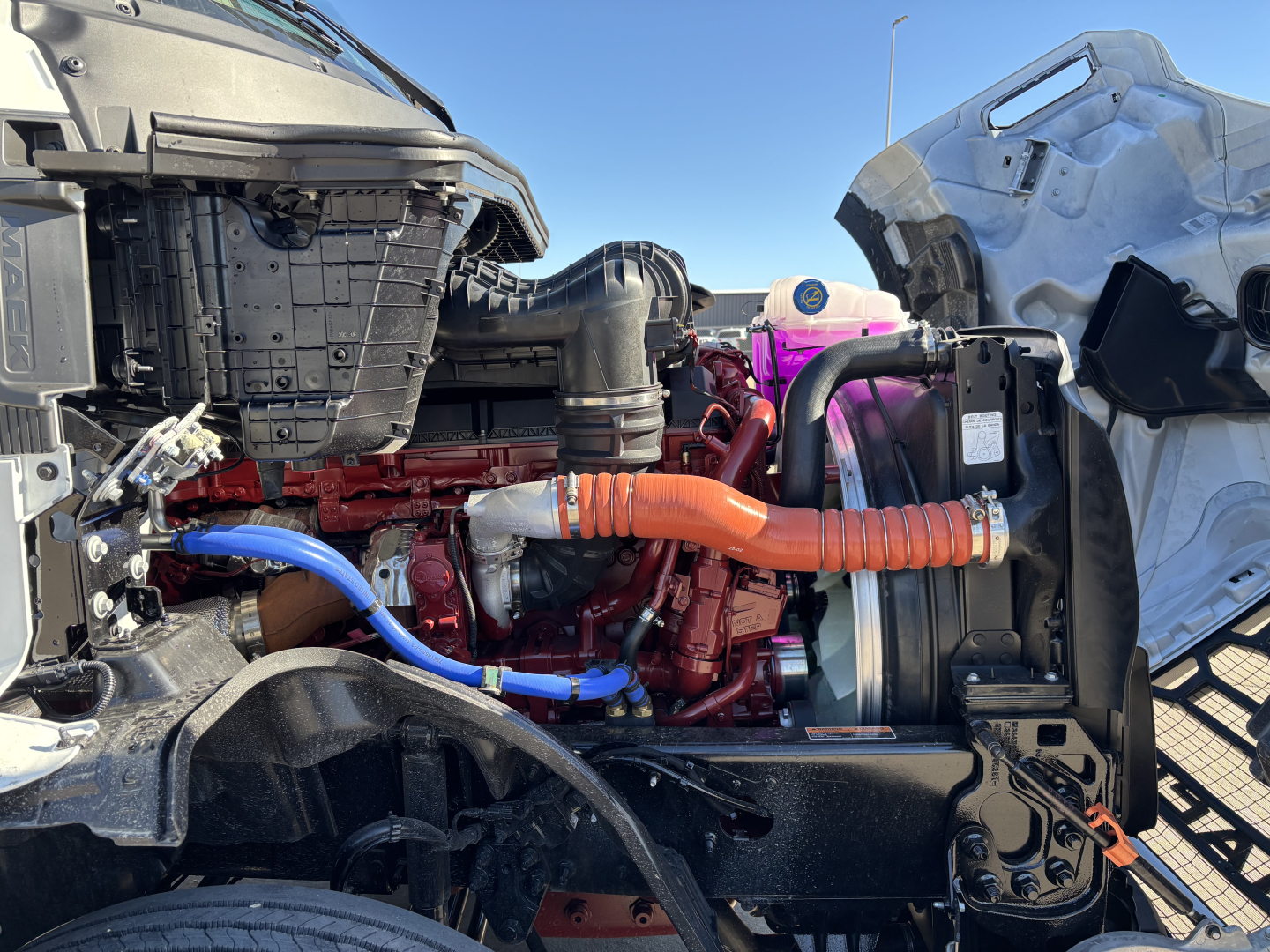 Mack PR64T engine bay with red engine, orange coolant hose, blue hose, and purple coolant reservoir; front cab area visible.