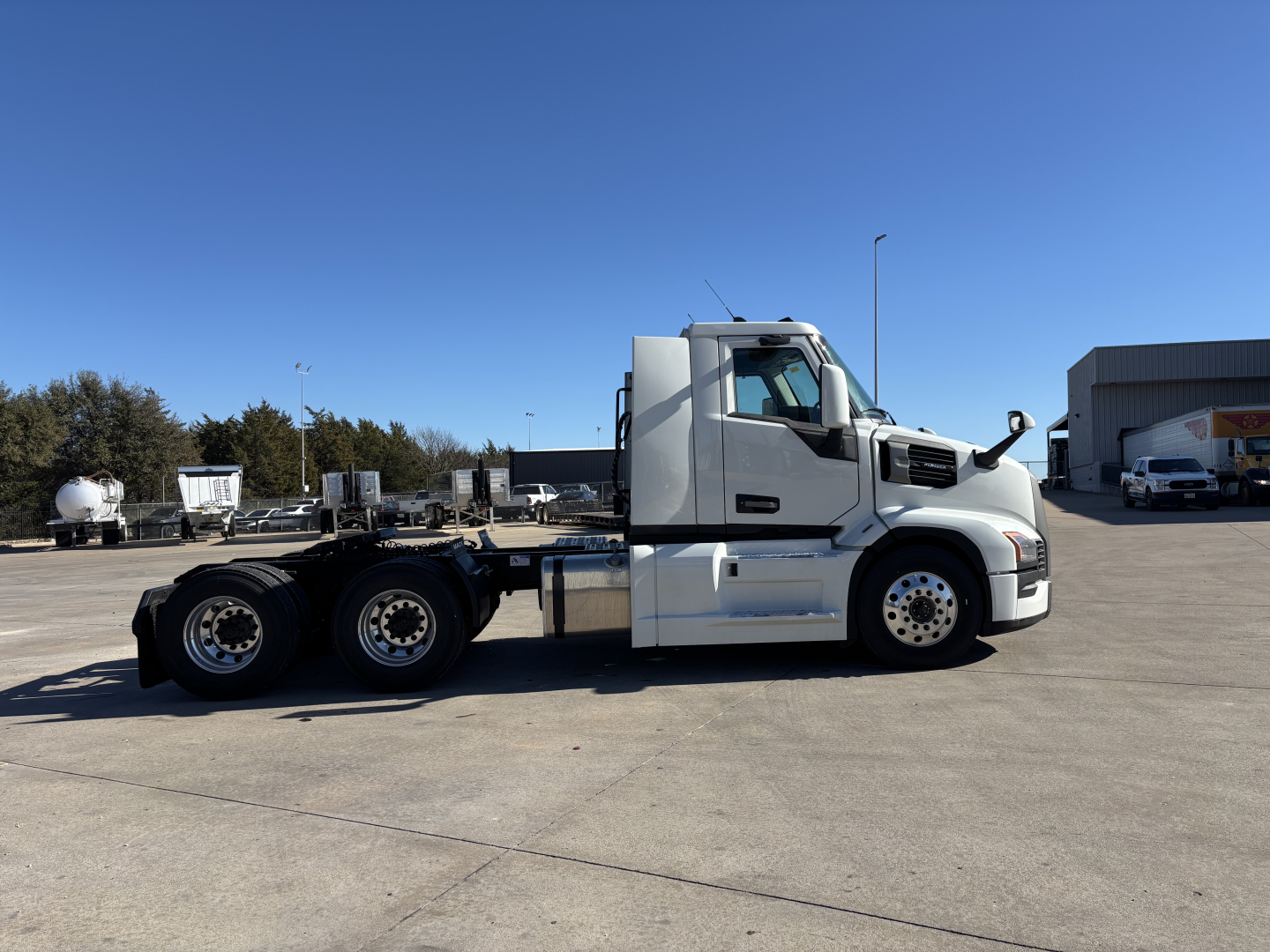 Side view of a white 2027 Mack PR64T truck chassis in a yard, with no trailer attached, showing dual rear axles and cab detail.