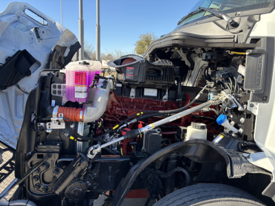 Truck engine bay open showing red engine block, hoses, reservoirs, and exposed components in a Mack PR64T cab; various fluid reservoirs visible.