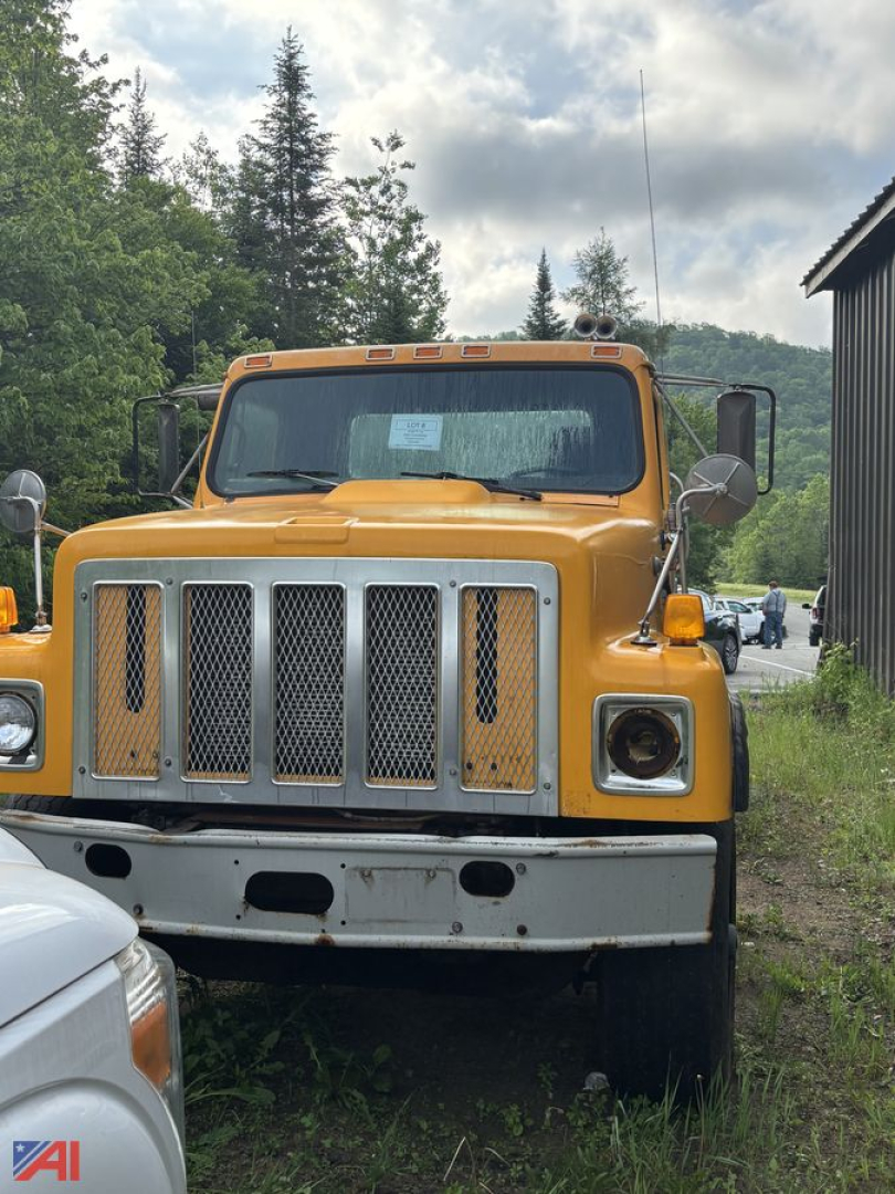 Front view of a yellow 1992 International S2574 Cab and Chassis truck with a large chrome grille and round headlights; overgrown grass at bumper; side mirrors; cloudy sky.