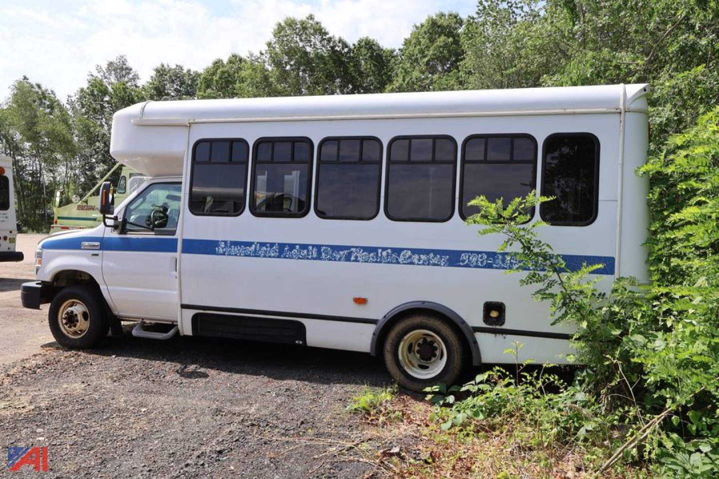 Ford E-350 shuttle bus with wheelchair lift, side windows, single blue stripe, rear dual wheels, parked on gravel with vegetation.