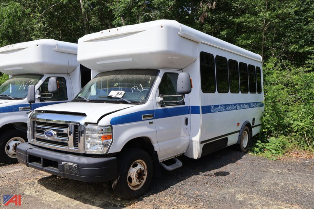 Ford E-350 wheelchair bus under a canopy showing front grille, headlamps, and passenger windows. Blue stripe along side and side door, no visible text legibly on bus.