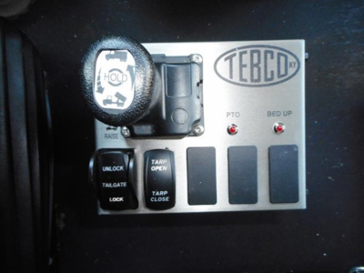 TEBCO KY control panel with hold lever and four switches labeled UNLOCK, TARP OPEN, TARP CLOSE, TAILGATE; plus LOCK, RAISE, and BED UP indicators; circular handle on left.