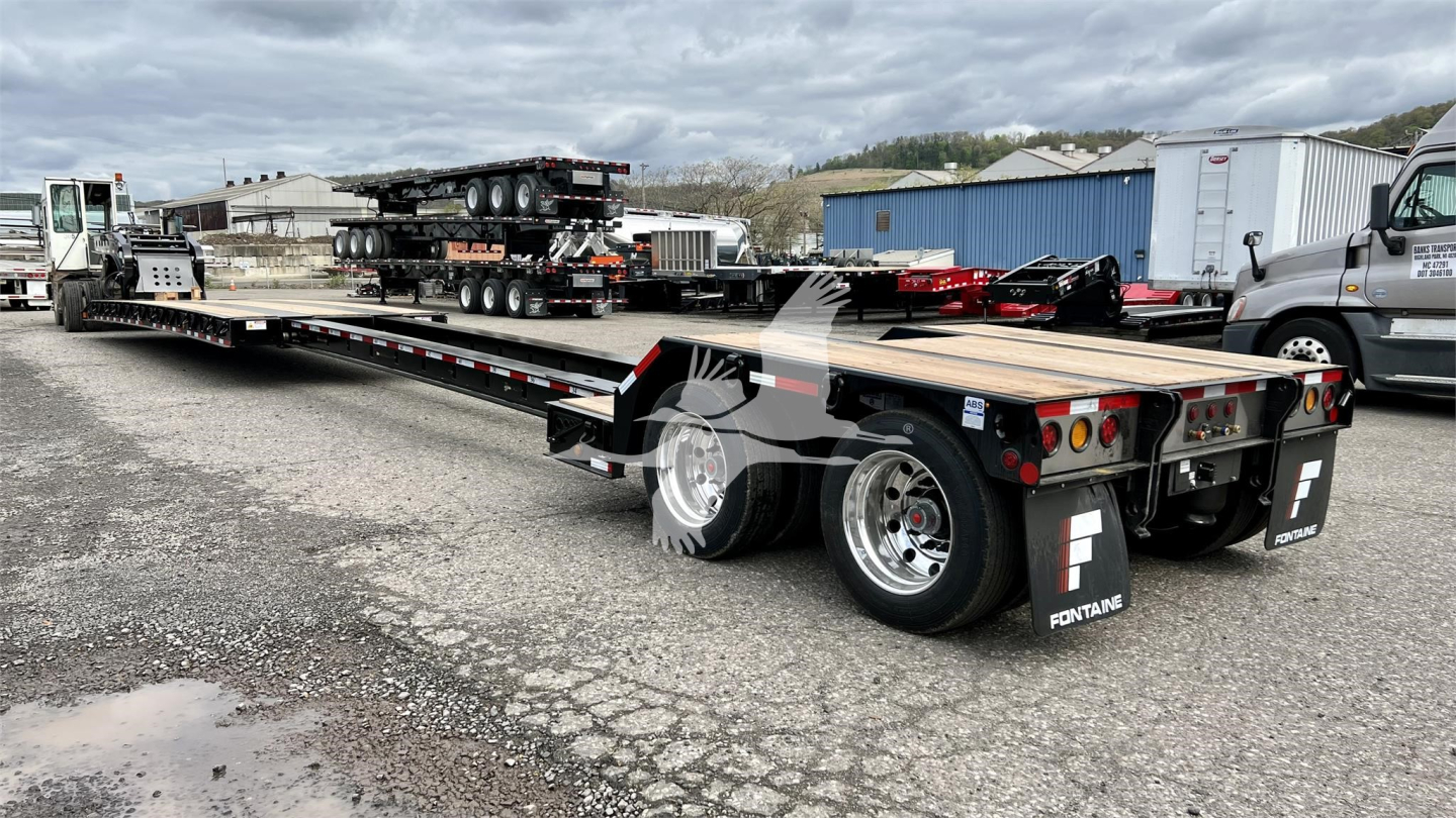 Lowboy trailer, Fontaine brand, long flatbed with dual rear axles showing storage boxes and lights; side rails and ramps visible, with a second trailer in background.