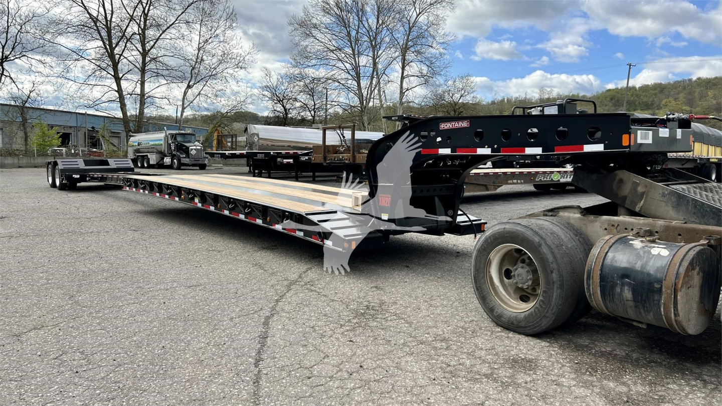 Large lowboy trailer with extended deck and black gooseneck, Fontaine XN20 logo visible, long steel frame, dual rear axle tires, equipment yard background.