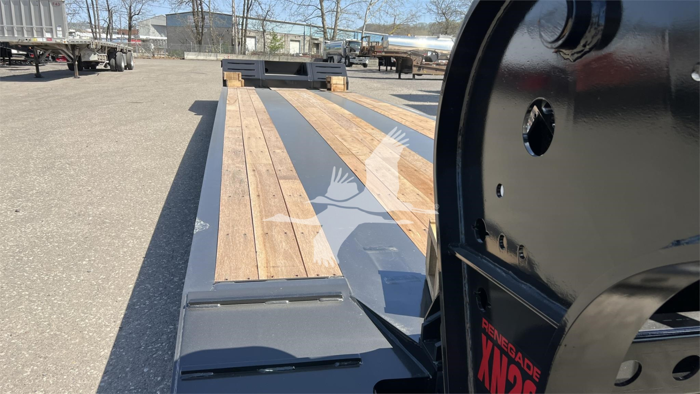 Lowboy trailer deck with wooden planks and black steel frame, close-up of neck area showing sunlit boards and highway lot in background.