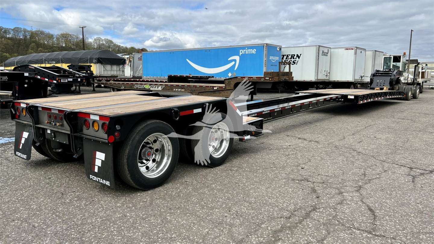 Lowboy trailer with multiple axles and chrome wheels showing Fontaine branding and rear view, long flatbed with other trailers in the background.