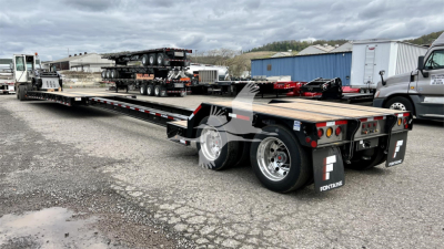 Lowboy trailer, Fontaine brand, long flatbed with dual rear axles showing storage boxes and lights; side rails and ramps visible, with a second trailer in background.