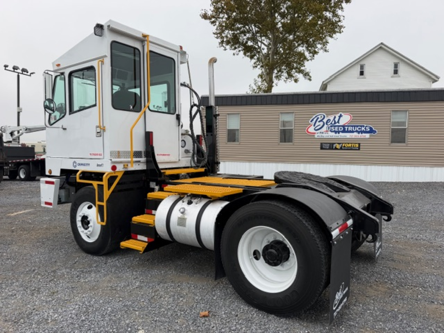 Truck chassis cab showing TJ5000; diesel fuel tank and rear tires visible; white cab with yellow accents and black fender; yard setting with Best Used Trucks sign.