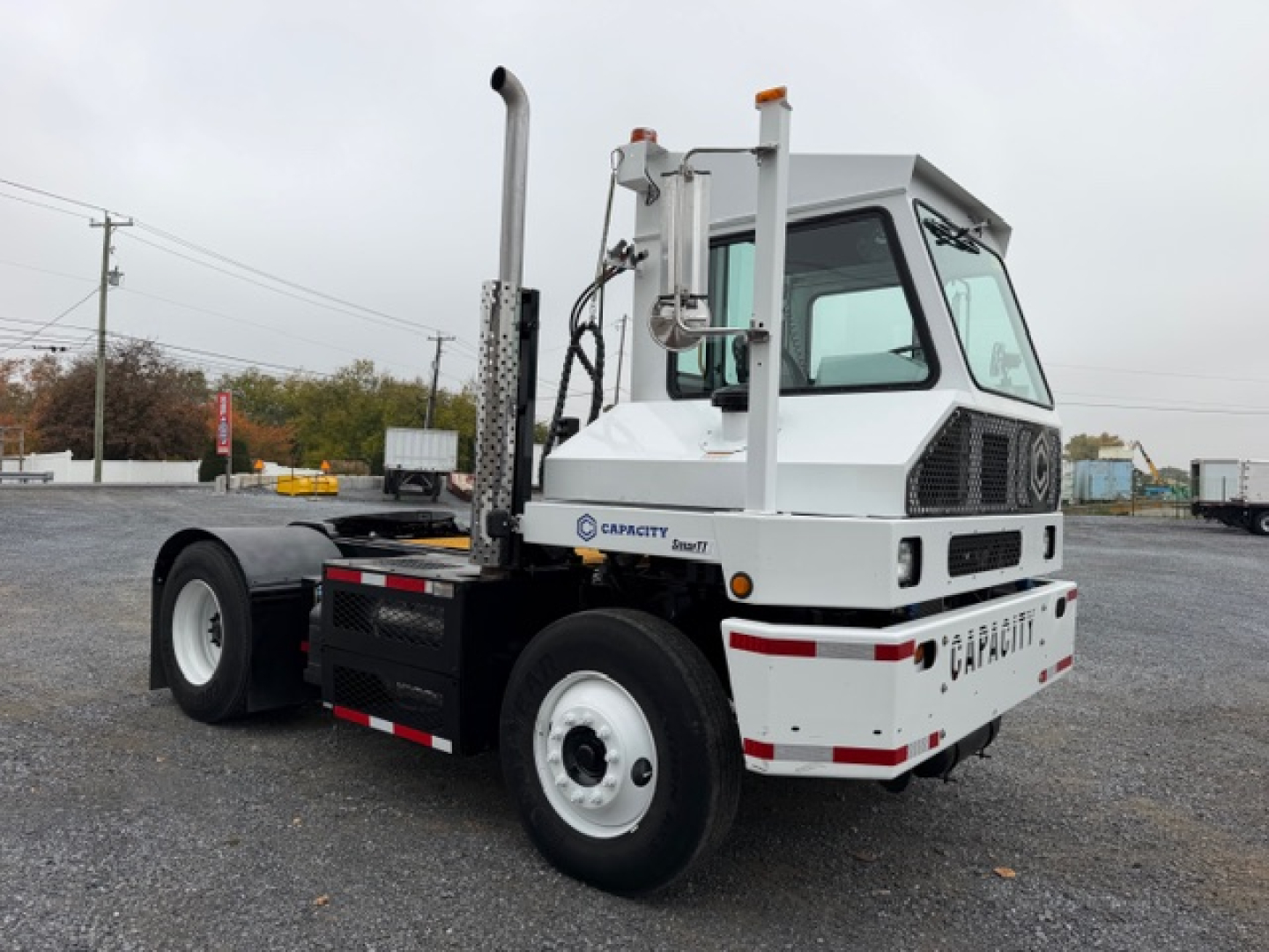 Capacity TJ5000 yard truck with white cab, dual exhaust stacks, and rear chassis; shows Capacity logo and model name on cab and rear bumper; metal framework and tires visible.