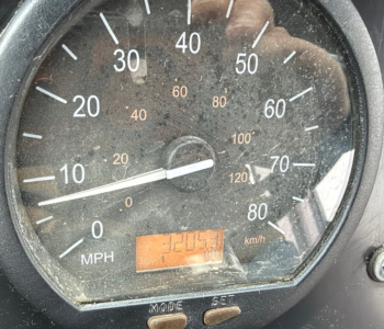 Gauge cluster showing speedometer with mph scale and an orange digital readout display. Dim instrument panel with numbers 0-80 km/h marks and reflection of a person taking the photo.