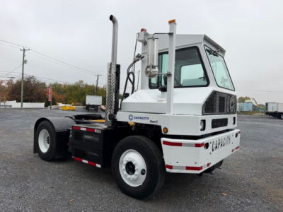 Capacity TJ5000 yard truck with white cab, dual exhaust stacks, and rear chassis; shows Capacity logo and model name on cab and rear bumper; metal framework and tires visible.