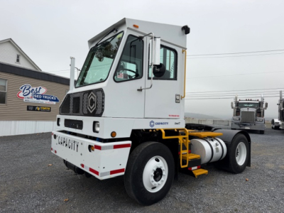 Truck cab of a TJ5000 capacity industrial tractor showing capacity logo on front and side, white cab with black grille, yellow step rails, fuel tank, and rear dual wheels.