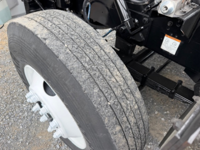 Truck wheel and front suspension area showing tire tread and wheel hub, partial chassis, and a sticker on the body.