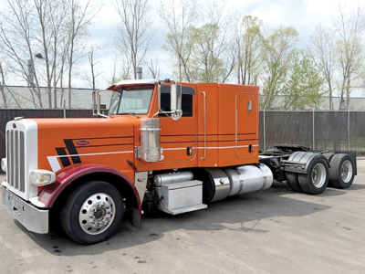 Peterbilt 388 tractor in orange with sleeper cab, chrome exhaust stacks, and dual rear axles; side view showing fuel tanks and steps.