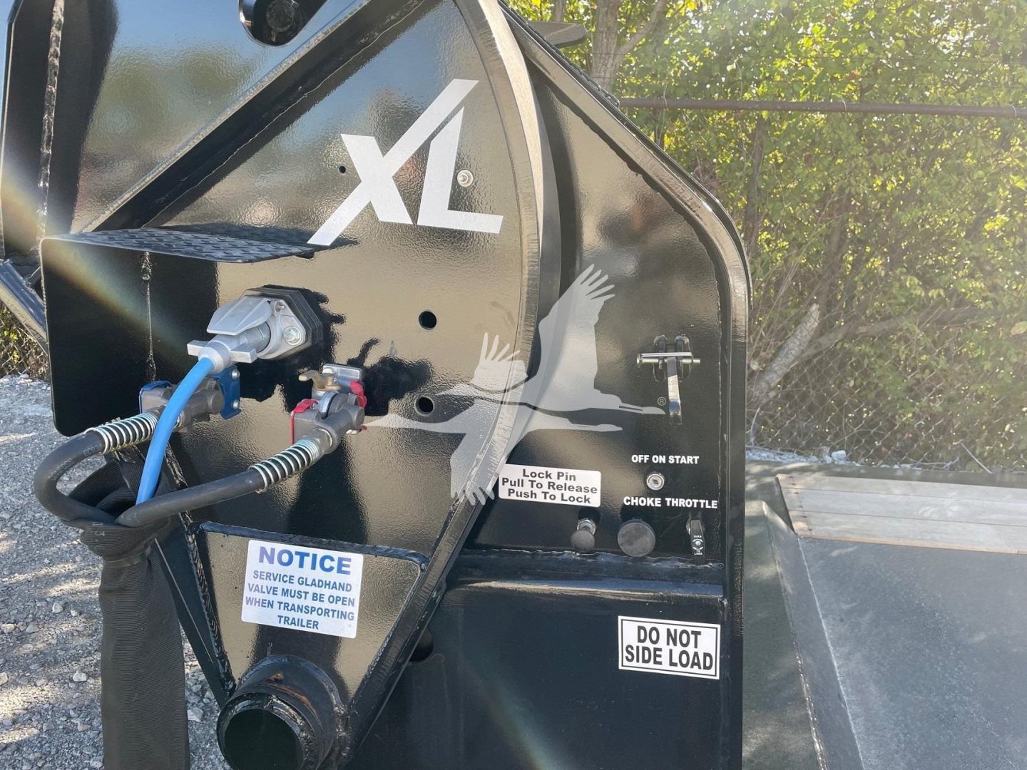 Compact trailer side with XL branding and hydraulic hoses connected to a coupling; visible labels include NOTICE, DO NOT SIDE LOAD, and CHOKE THROTTLE on the black trailer body.