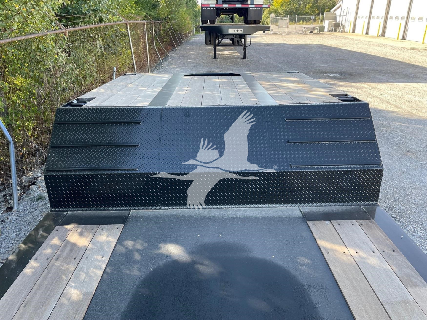Deck truck trailer with a raised black metal toolbox and a white bird logo on the front; wooden deck planks and a vehicle in the distance.