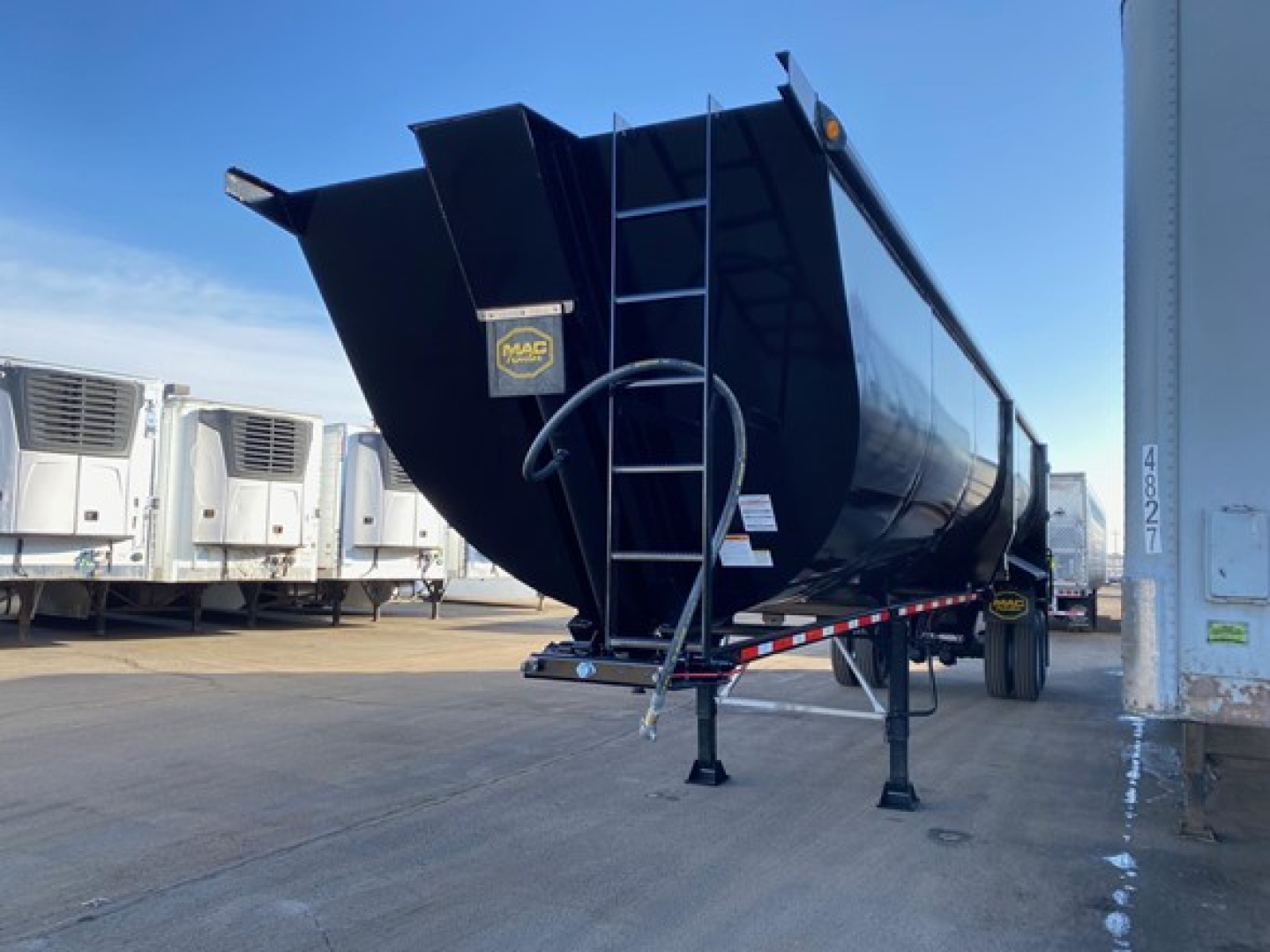 Dump trailer with black glossy finish showing Mac Trailers logo on front panel and ladder, front legs deployed, side rails, and attached hitch.