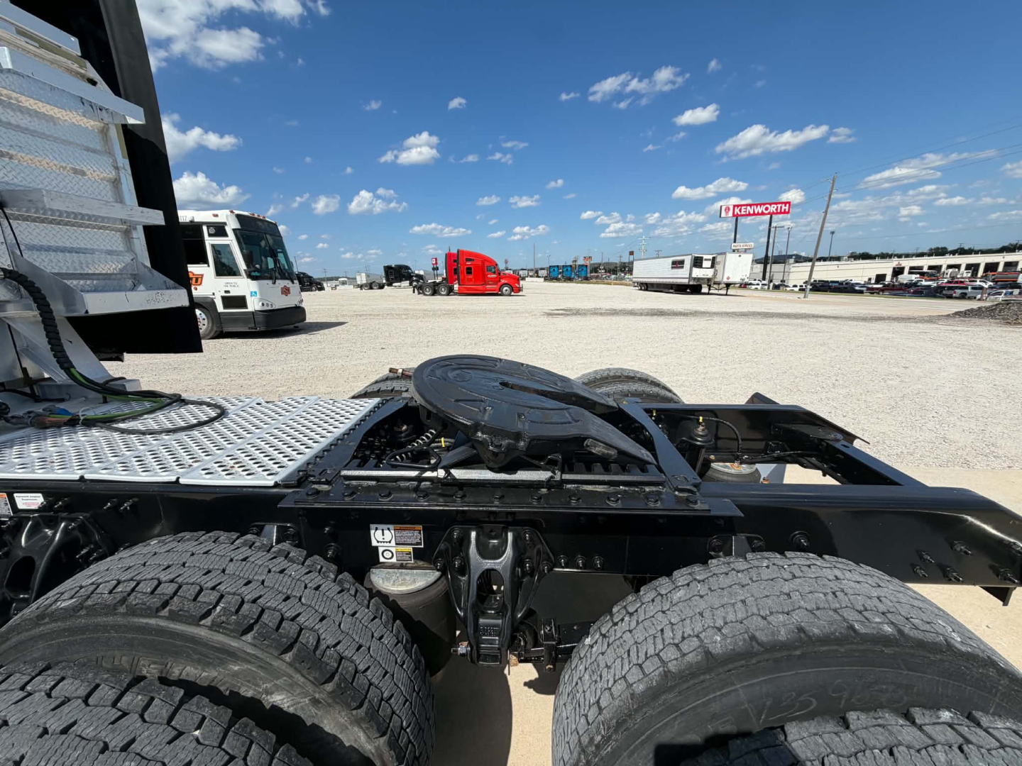 Kenworth T680 truck chassis view showing dual rear axles and fifth wheel mounting area with nearby white and red trucks in yard.