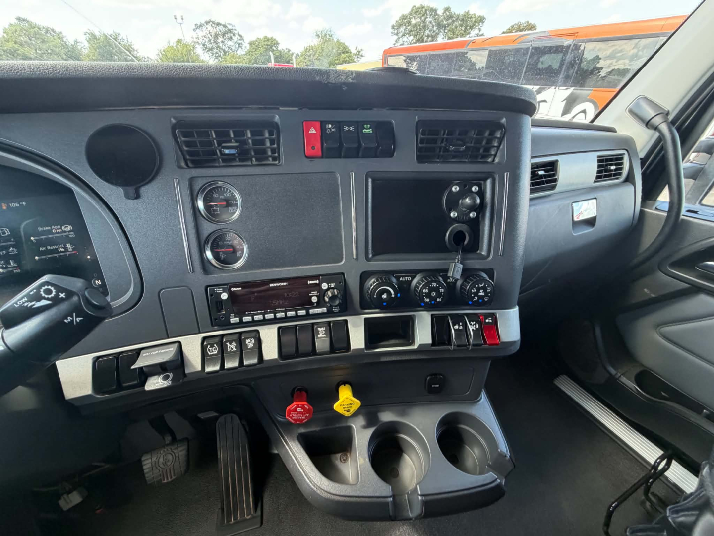 Kenworth T680 dashboard/driver area showing center console with vents, hazard light, various switches, gauge cluster, and cup holders. Text visible includes: none clearly legible except small labels on switches.