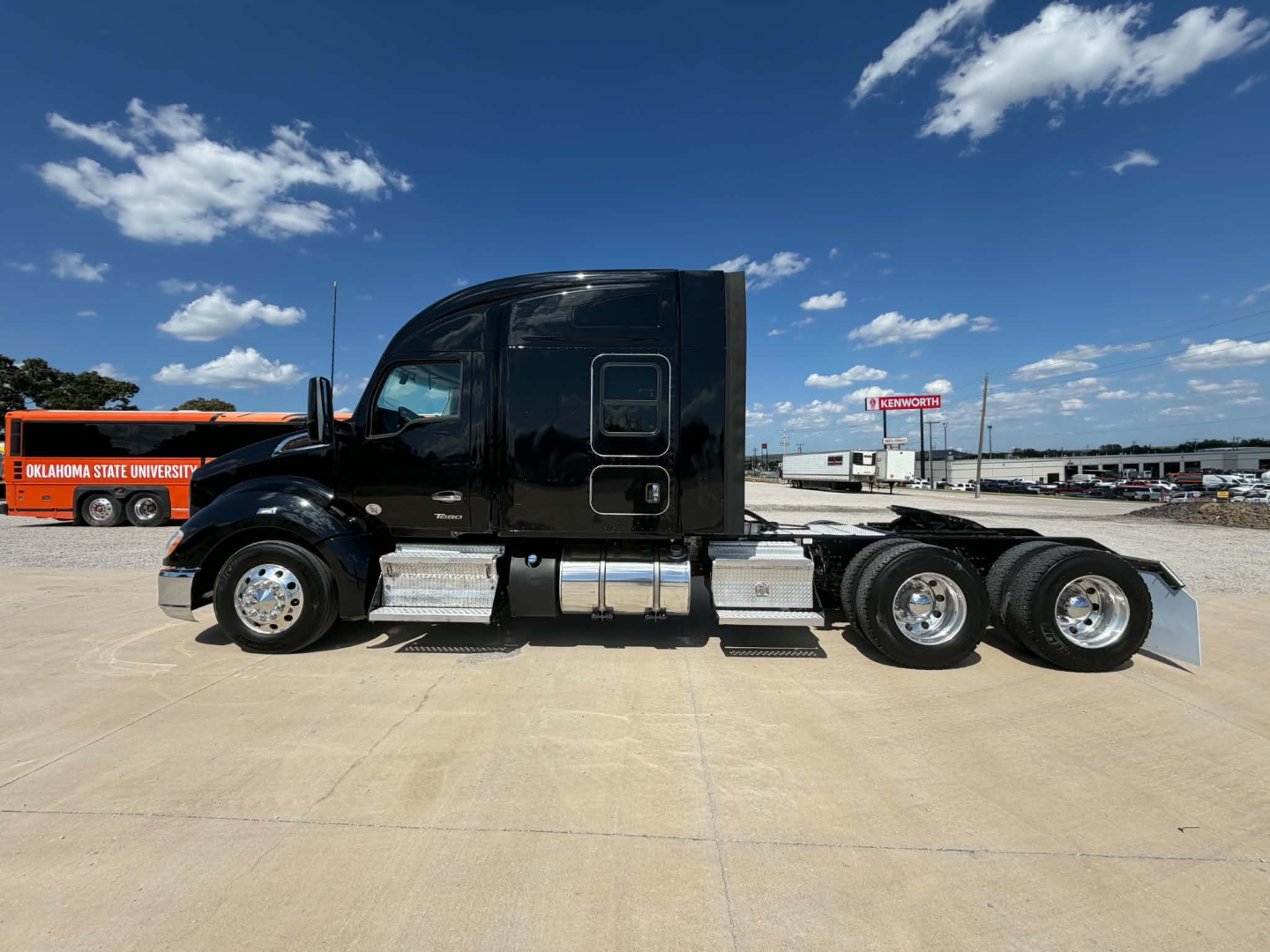 Side view of a black 2022 Kenworth T680 tractor with streamlined sleeper cab, chrome exhaust, and three rear axles on a concrete lot.