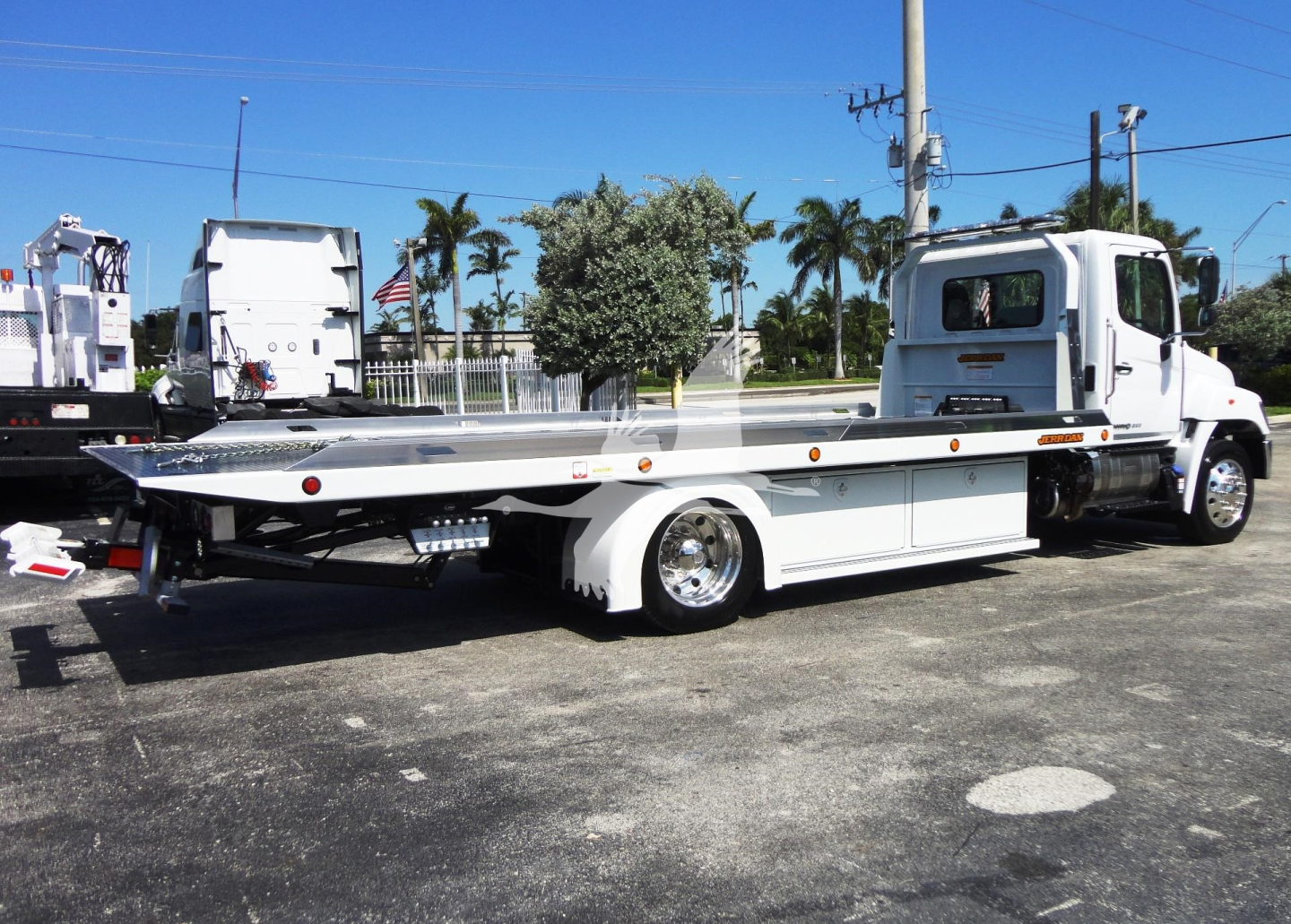 Flatbed tow truck in side view with white cab and long aluminum deck, rollback equipment visible.