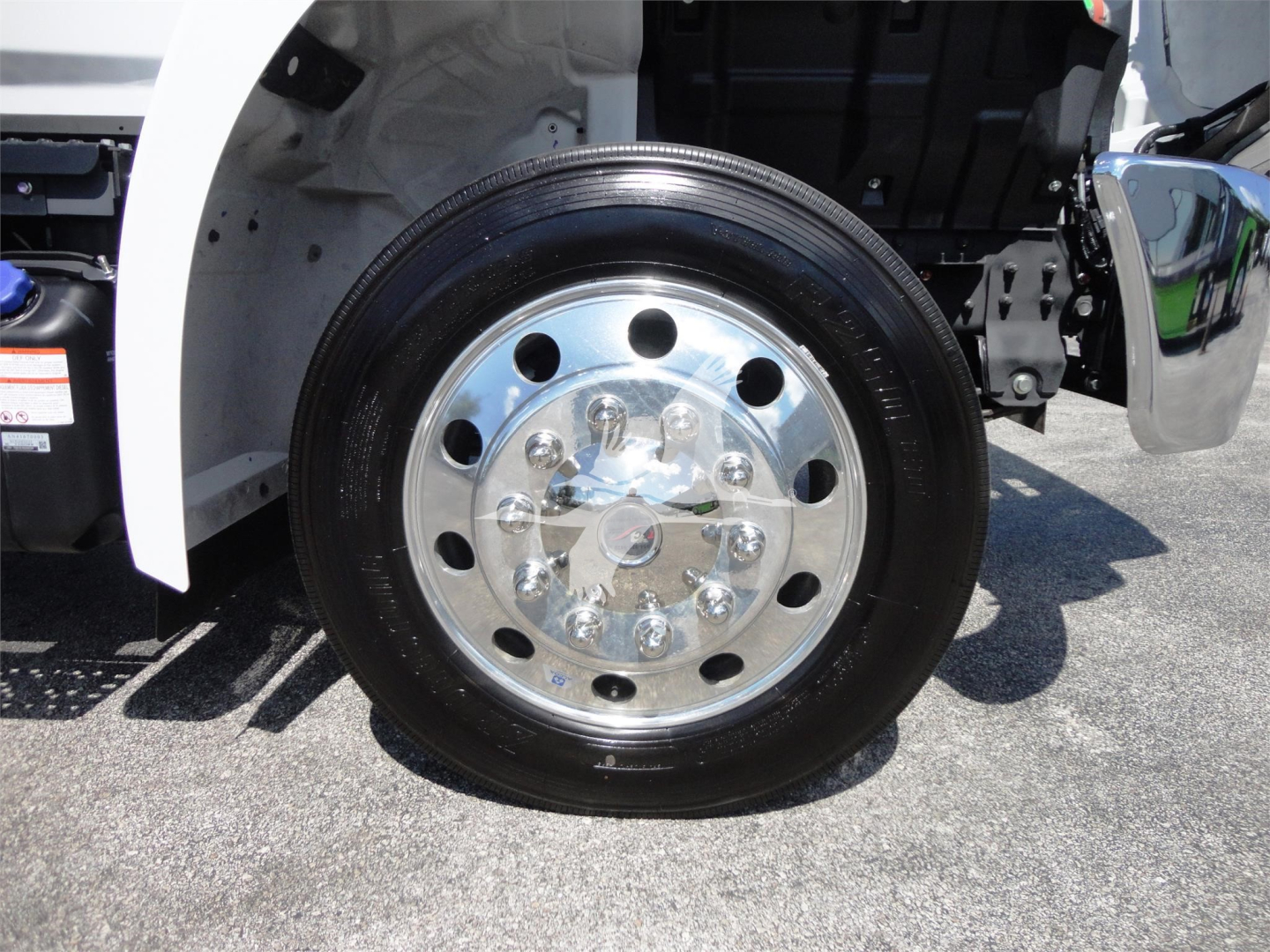 Front wheel of a 2022 Hino 268A rollback truck showing shiny chrome hub and black tire with wheel lug nuts; partial side of vehicle and fender visible.
